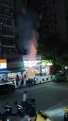 An electric scooter is throwing a large and violent flame upwards while parked in front of some stores. A man topples it over in an attempt to extinguish the flames